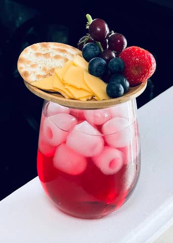 Glass of iced pink drink topped with a bamboo lid holding cheese slices, grapes, blueberries, strawberries, and a cracker