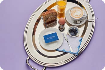 Breakfast tray with coffee, orange juice, sliced bread, an egg, blueberries, and a blank card on a white plate with a blue napkin and spoon