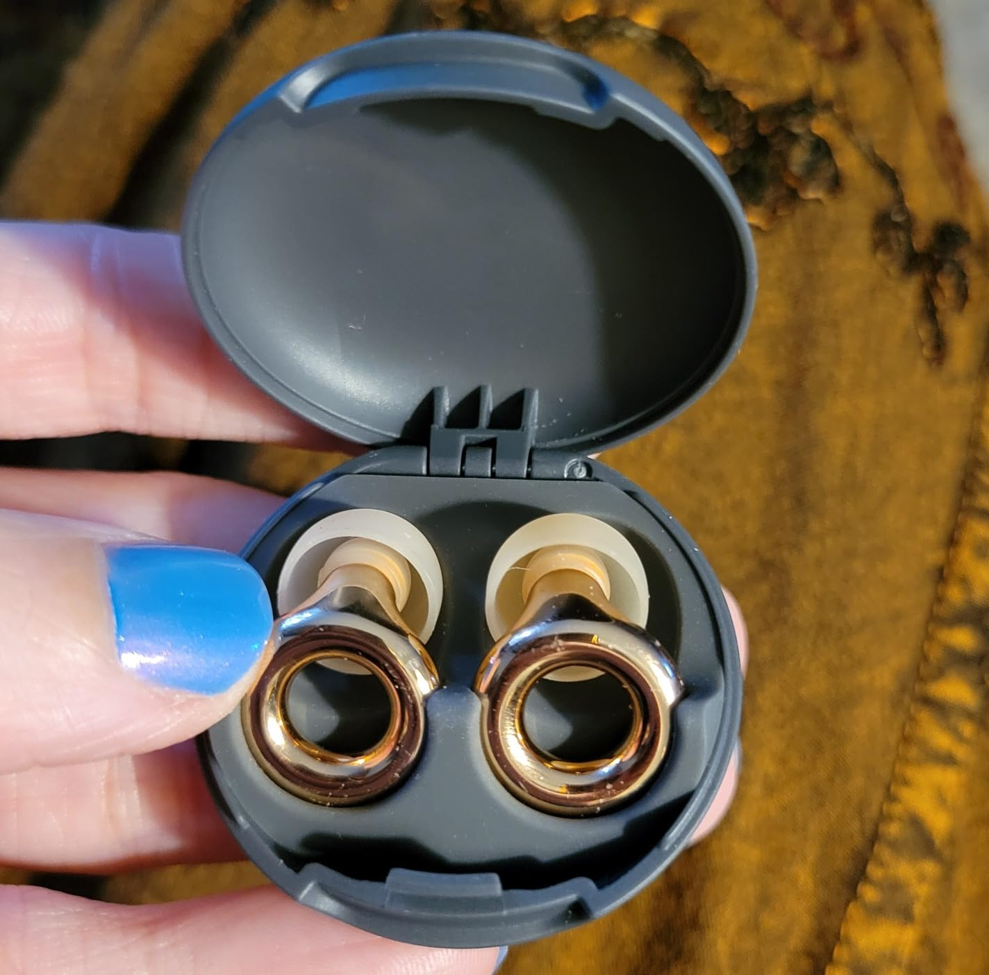 Open case displaying wireless earbuds with metal loop design. Person holding the case has blue-painted nails