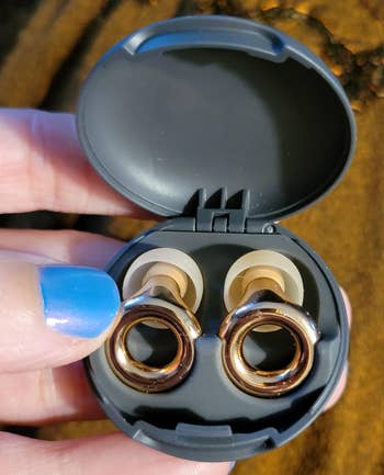 Open case displaying wireless earbuds with metal loop design. Person holding the case has blue-painted nails