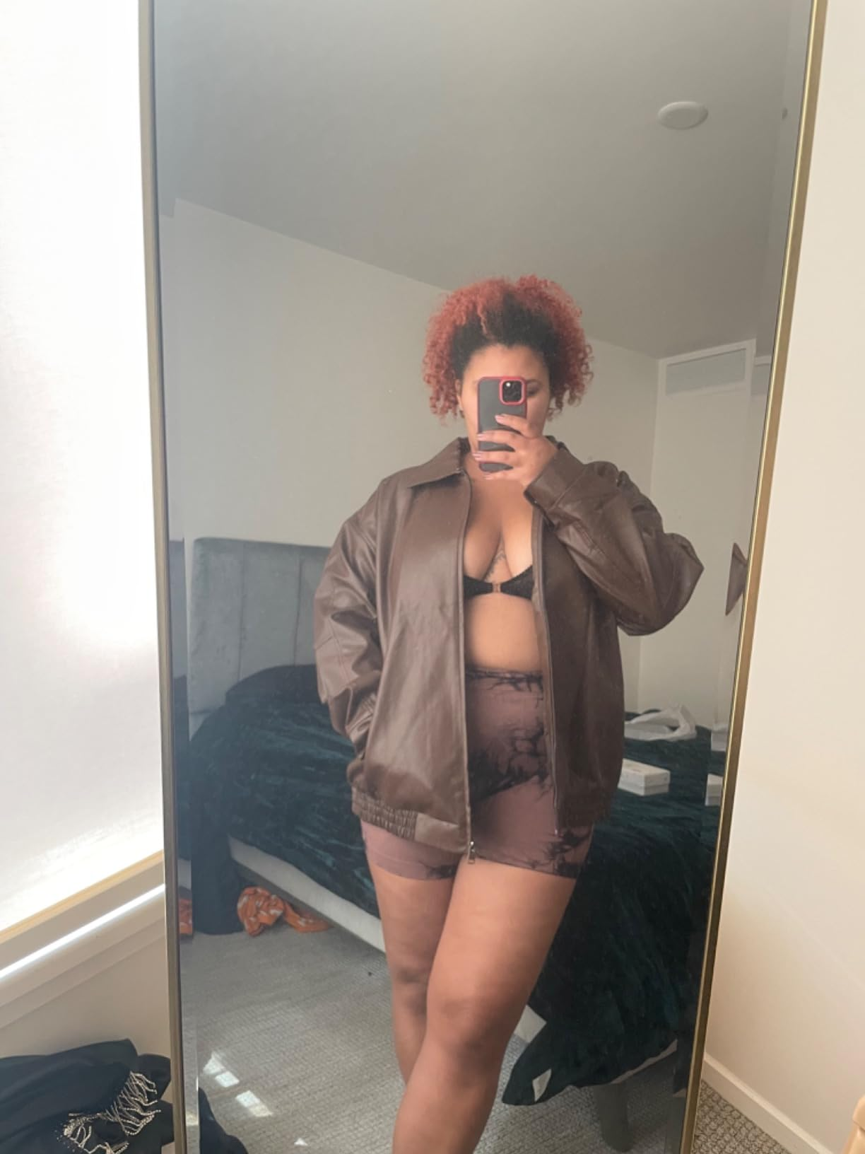 Person successful  a brownish  leather jacket, achromatic  bra, and patterned shorts, taking a reflector  selfie successful  a bedroom