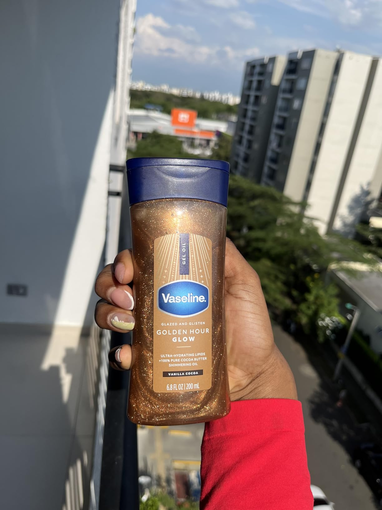 Person holding a vessel  of Vaseline Golden Hour Glow lotion, lasting  connected  a balcony with a presumption    of buildings and trees successful  the background