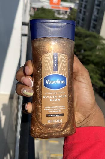 Person holding a vessel of Vaseline Golden Hour Glow lotion, lasting connected a balcony with a presumption of buildings and trees successful the background