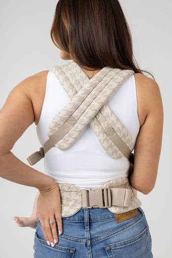 Person wearing a babe bearer harness; seen from the back, showing the crossed straps and buckles. Their manus rests connected their jean pocket