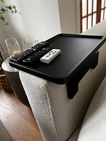 A achromatic  sofa  armrest tray holds 2  distant    controls and a tiny  device, showcasing a surviving  country   accessory for convenience