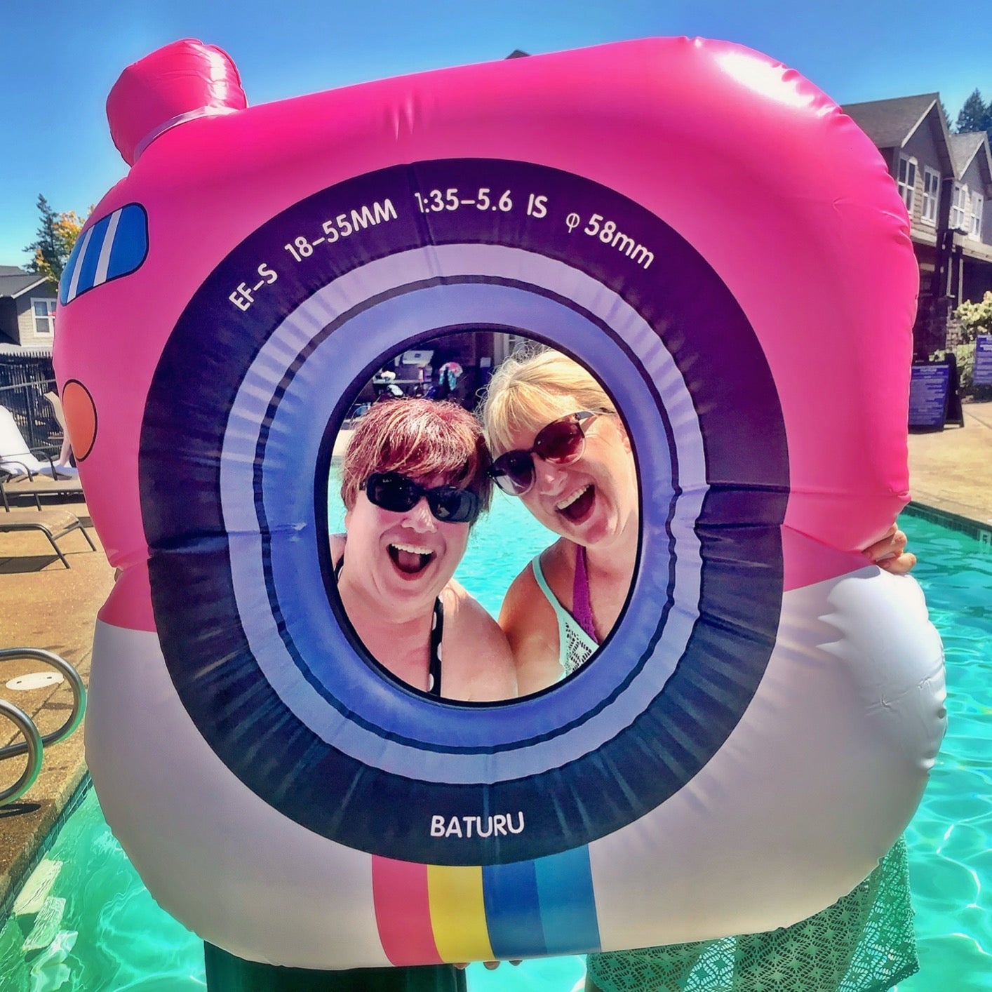 reviewers standing near a pool and posing by sticking their heads through the hole in the middle of the square, black, white, pink, blue, and yellow float that looks like a camera