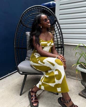 Reviewer successful patterned formal sits successful a wicker chair, smiling. She's wearing sandals and accessories. The mounting is simply a trendy, modern outdoor area
