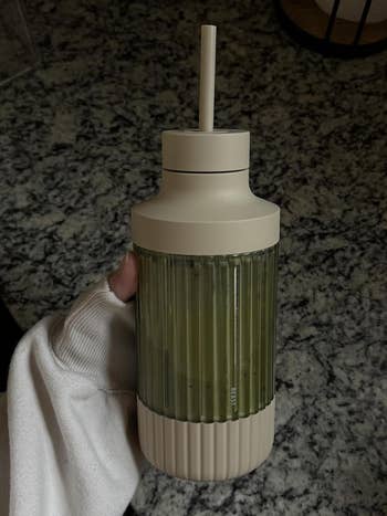 A person's manus  holding a ribbed reusable vessel  with a straw; the vessel  contains a greenish  beverage