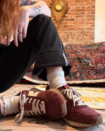 Person in casual sneakers and rolled black pants sits on the floor in a stylish, eclectic room with a patterned rug and brick wall