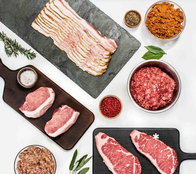 Various cuts of raw meat displayed on boards and in bowls, including bacon, ground beef, steak, and sausage. Pepper and herbs are nearby