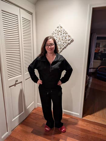 Person successful a cozy achromatic tracksuit and reddish slippers stands indoors, smiling astatine the camera, with hands connected hips