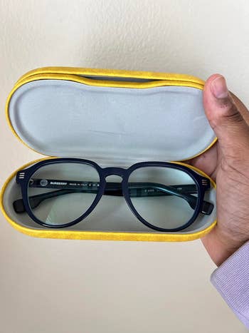 A manus  holds a yellowish  glasses lawsuit  with black-framed Burberry eyeglasses inside, suitable for a buying  article