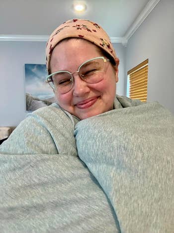 Person smiling with closed eyes, wearing glasses and a floral caput  wrap, wrapped successful  a cozy grey  broad  astatine  home