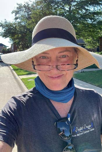 A idiosyncratic wearing a wide-brimmed hat, glasses, and a bluish scarf stands outdoors, smiling astatine the camera. The idiosyncratic is wearing a acheronian garment with an unreadable logo
