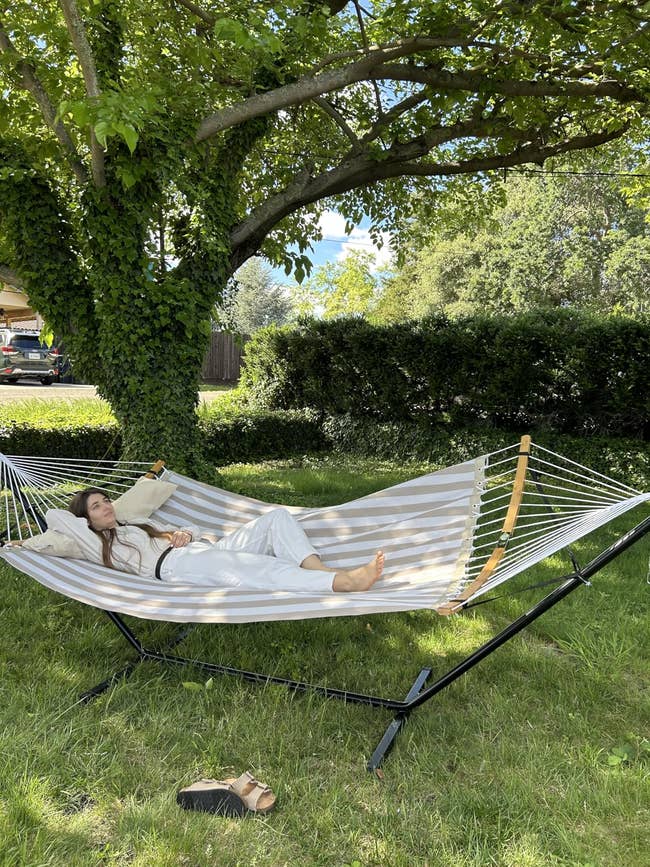 Person relaxing connected  a striped hammock nether  a histrion   successful  a garden. They're wearing a light, casual outfit. Nearby, a brace  of sandals is connected  the grass
