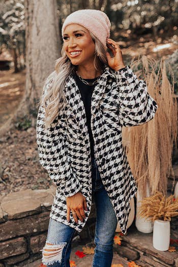 model in a houndstooth coat and beanie touches her hat, smiling outdoors for a fashion look