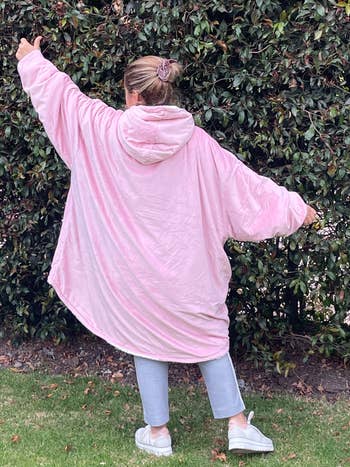 Person outdoors wearing oversized hooded blanket-style loungewear, facing away, arms outstretched. Comfortable and casual look