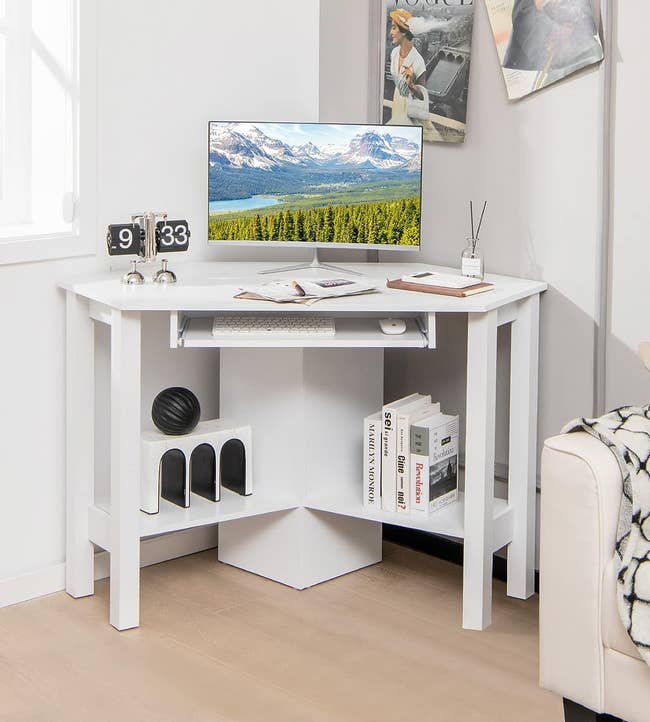 White wood triangle desk with a keyboard drawer and shelves under with books, in the corner of a room