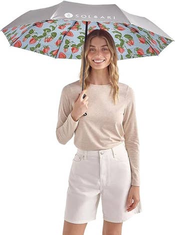 Smiling idiosyncratic holds an umbrella with a strawberry pattern, wearing a casual long-sleeve apical and shorts