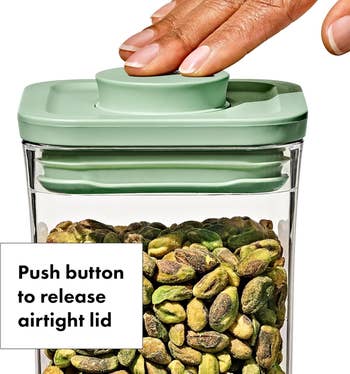 Hand pressing button on a clear container lid, releasing the airtight seal, filled with pistachios. Text:
