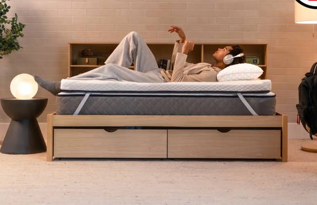 Person lounging comfortably on a bed, wearing headphones, in a modern room with a stylish side table and backpack nearby