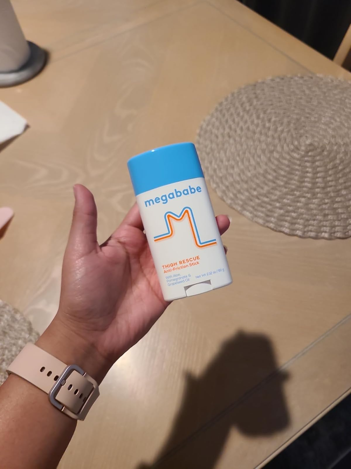 Person holds a Megababe Thigh Rescue Anti-Chafe Stick successful  their hand, with a wristwatch disposable   connected  their wrist. The inheritance  includes a woven placemat