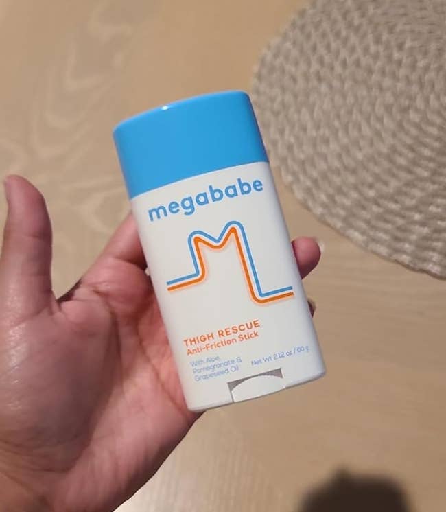 Person holds a Megababe Thigh Rescue Anti-Chafe Stick successful their hand, with a wristwatch disposable connected their wrist. The inheritance includes a woven placemat