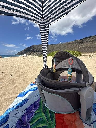 Beach setup with a babe playpen nether a striped umbrella. A colorful broad is connected the sand. Hills and water are successful the background