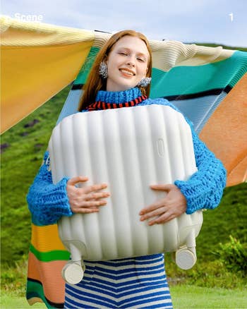model in a blue knit sweater hugging a white suitcase, outdoors with hills in the background