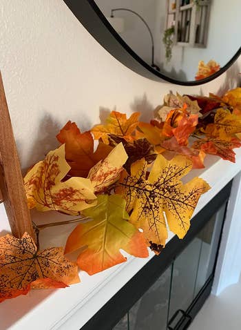 Decorative autumn-themed garland with assorted artificial leaves displayed connected  a mantel nether  a circular  mirror