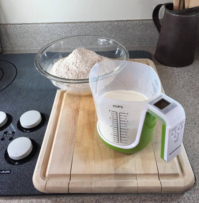 A bowl of flour and a measuring cup of milk on a wooden cutting board next to a stove