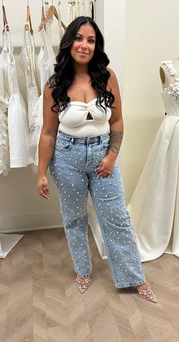 Person successful pearl-embellished jeans and achromatic apical stands successful a bridal boutique adjacent to a mannequin wearing an elegant achromatic wedding dress