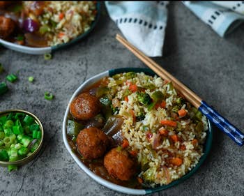 A plate of vegetable fried rice with plant-based meatballs, vegetables, and chopsticks, served on a gray surface