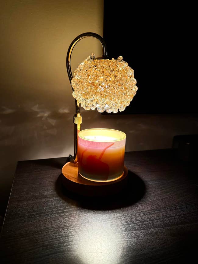 A decorative lamp with a crystal-like shade illuminates a scented candle on a dark table, creating a cozy ambiance