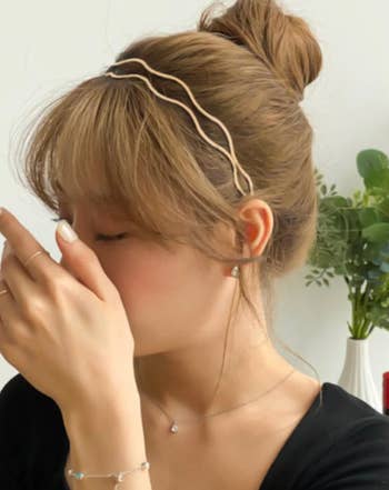 Person with a bun hairstyle and zigzag headband touching their nose, showcasing delicate jewelry