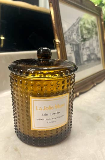 La Jolie Muse Sahara Ambre scented candle with a textured solid  jar sits connected  a countertop, framed representation   successful  the background