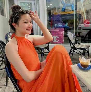 Woman sitting at a cafe table, wearing a sleeveless orange dress, with a hand in her hair