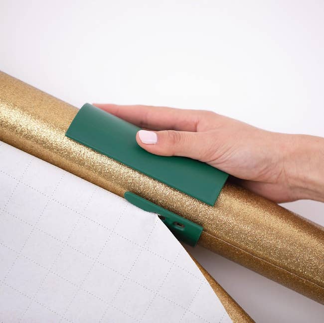 Hand using a green tool to cut gold glitter wrapping paper on a grid-lined surface, ideal for precise gift-wrapping