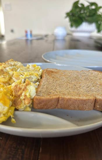 Scrambled eggs and toast on a white plate on a wooden table, with a plant in the background