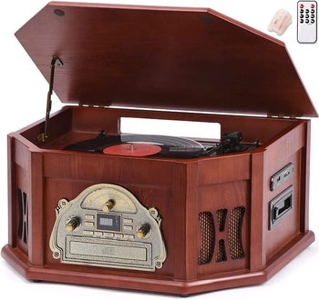 Brown wooden antique-styled turntable with matching lid open and black vinyl inside; front-view with silver plate and adjustable dials underneath silver remote on a white background