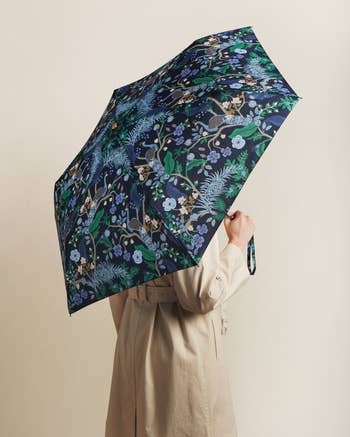 Person holding an umbrella with a floral pattern, obscuring their face, dressed in a beige coat