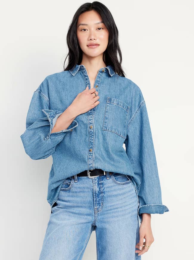 Person wearing a denim garment  and jeans, posing with 1  manus  adjusting the collar. Casual and relaxed style