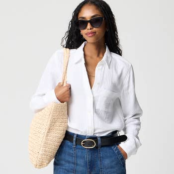 Model in a casual chic outfit with sunglasses, white blouse, denim skirt, and straw tote
