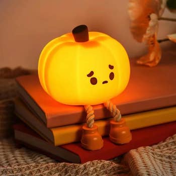 Cute pumpkin-shaped night light with a sad face, sitting on stacked books, featuring rope legs and shoes. Cozy and whimsical décor piece perfect for autumn