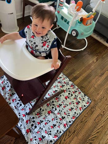 Baby in high chair with Mickey Mouse bib and matching floor mat. Perfect for Disney-loving families