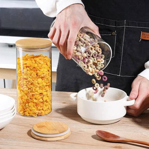 Person pouring cereal into a vessel  adjacent  to a jar of cornflakes connected  a woody  counter