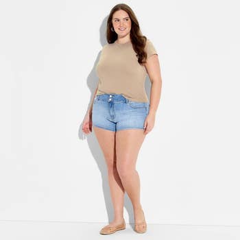Person wearing a casual outfit with a beige top, denim shorts, and loafers, posing against a plain background