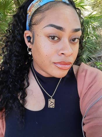 A person wearing a headband, earbuds, black top, and pink jacket; accessorized with a necklace featuring a rectangular pendant, poses outdoors