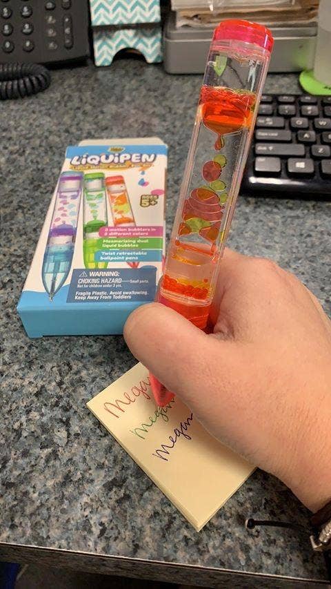 A manus holds a liquid-filled pen connected a table with colorful notes. A packaging container for the pen is besides disposable adjacent to a machine keyboard
