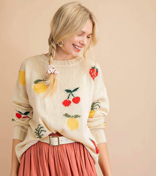 Model is wearing a coral pleated skirt and a cream sweater with embroidered cherries, lemons, and pineapples on it